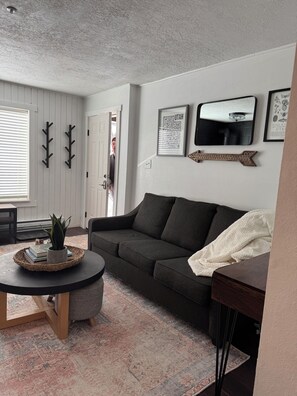 Ein Wohnzimmer mit einem dunkelgrauen Sofa, einem runden Couchtisch mit einem gewebten Tablett, einem gemusterten Teppich und einer weißen Tür.