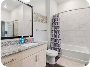 A bathroom with a white toilet, a sink with a mirror, a shower with a patterned curtain, and a tiled wall. - Modern & Lavish luxury 1 bedroom/1 bath in Deep Ellum Downtown Dallas (Dallas)