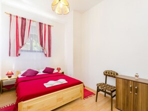 Um quarto com cama de madeira, roupa de cama vermelha, uma janela com cortinas vermelhas, abajur, mesa de cabeceira de madeira, guarda-roupa de madeira e uma cadeira estampada.