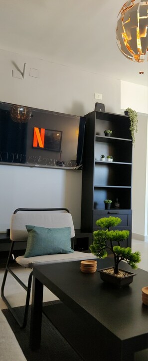 A modern living room with a sofa, a coffee table, a bonsai tree, a black bookshelf, and a wall-mounted TV with a Netflix logo. - Amazing Suites 2-bedroom apartment with AC in Punta Cana (Punta Cana)