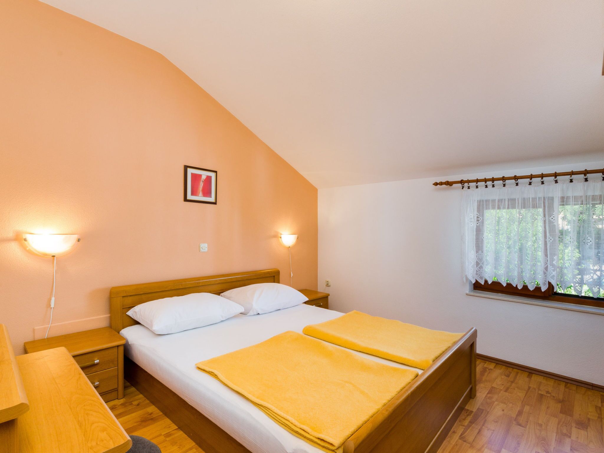 Un dormitorio con cama, lámparas de noche, una ventana con cortina y un cuadro en la pared.