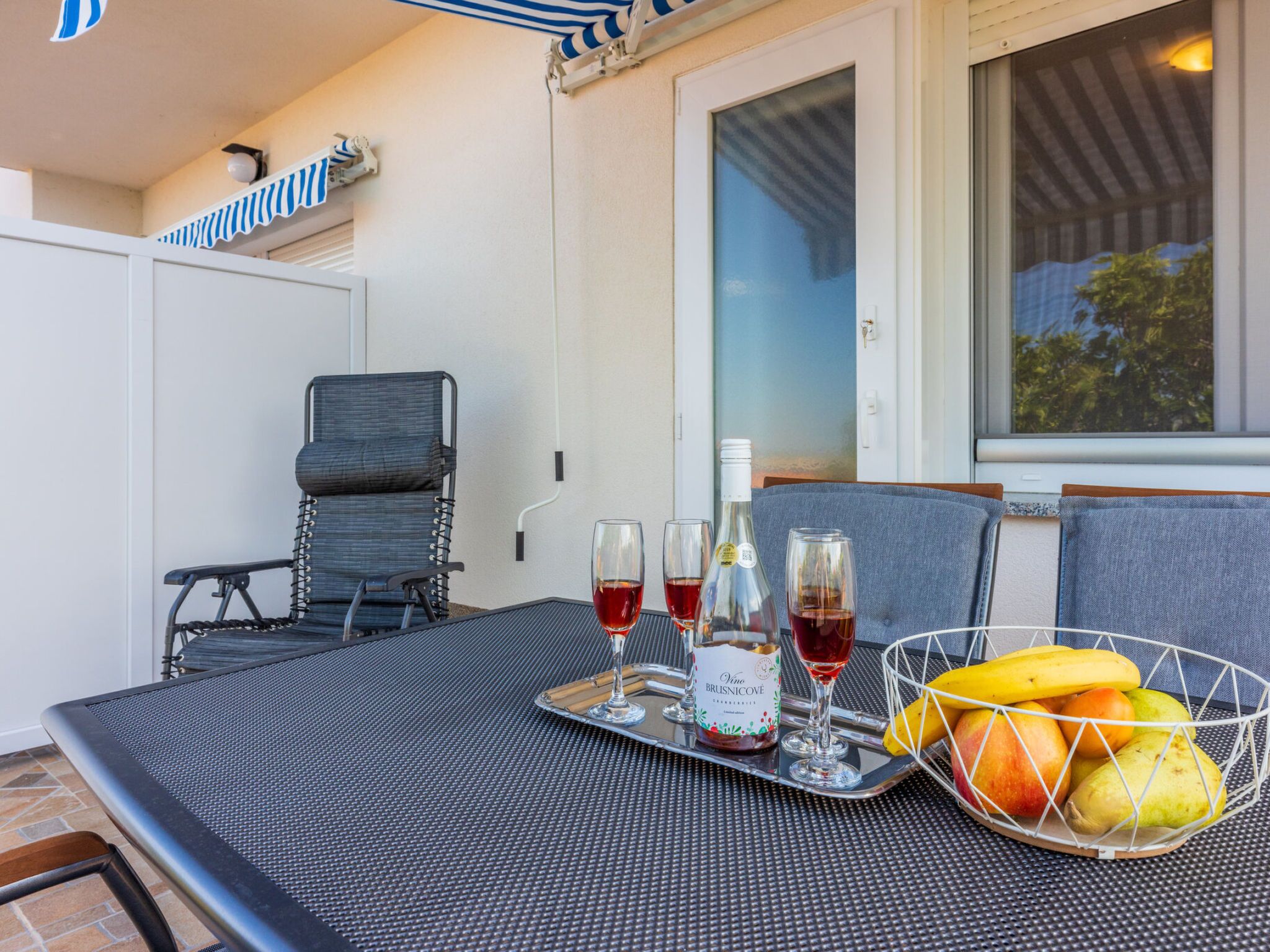 A patio with a table set for two, featuring a bottle of wine, glasses, and a fruit basket.
