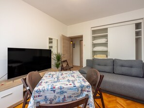 Ein Wohnzimmer mit grauem Sofa, Fernseher, Esstisch mit Stühlen und Bücherregal.