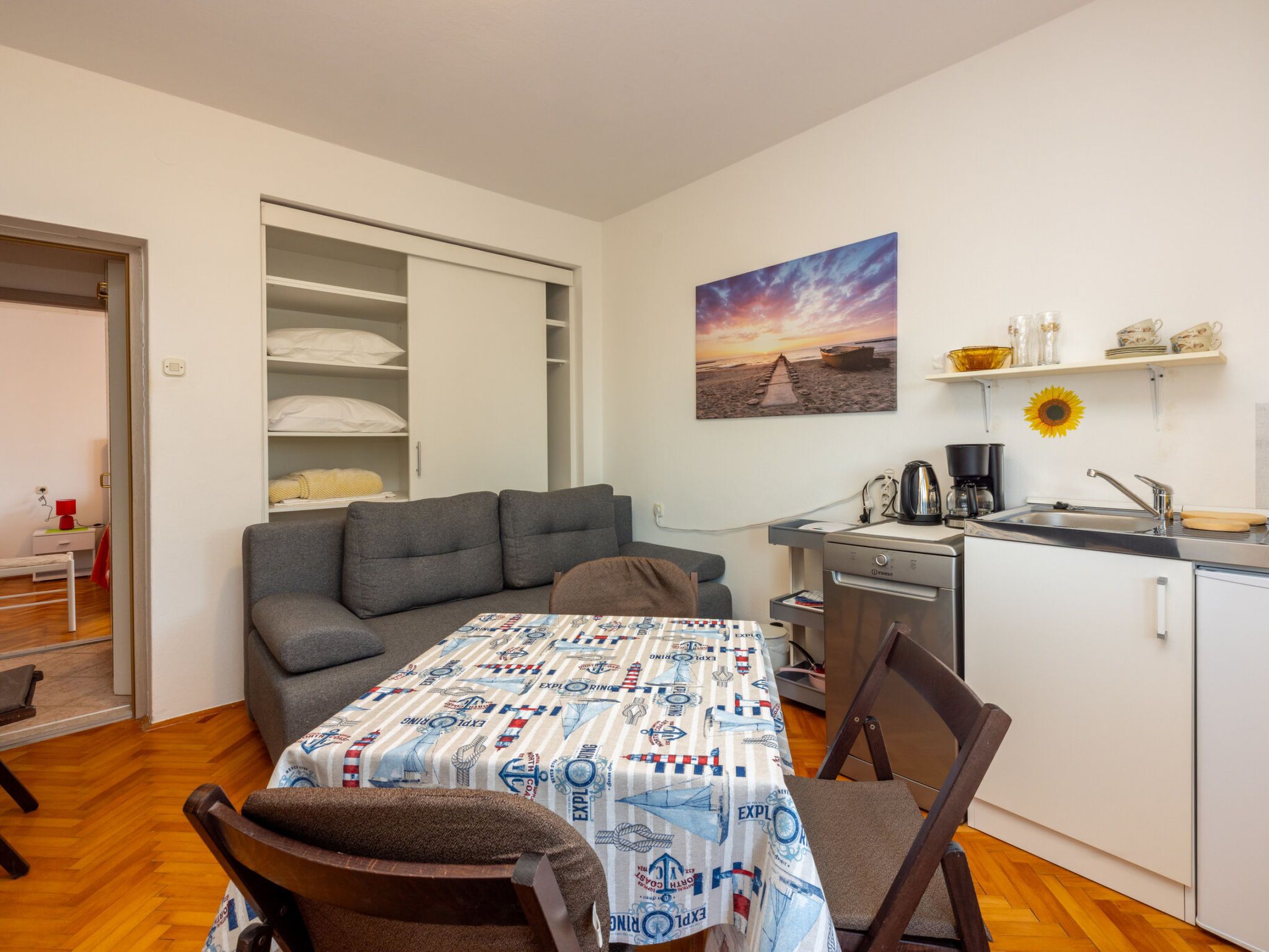 A compact room with a sofa, a dining table with a patterned tablecloth, a kitchenette, and a painting of a sunset.