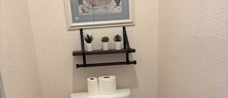 A bathroom with a toilet, a framed picture above the sink, and a shelf with potted plants.
