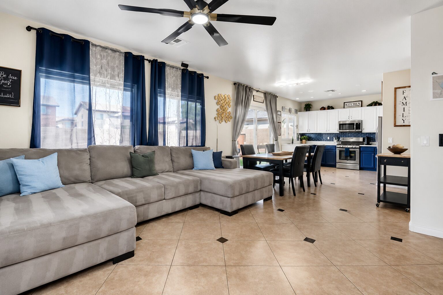 A modern living room with a sectional sofa, a dining area with a table and chairs, and a kitchen with blue cabinets and a microwave.