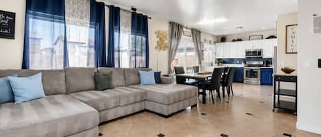 A modern living room with a sectional sofa, a dining area with a table and chairs, and a kitchen with blue cabinets and a microwave.