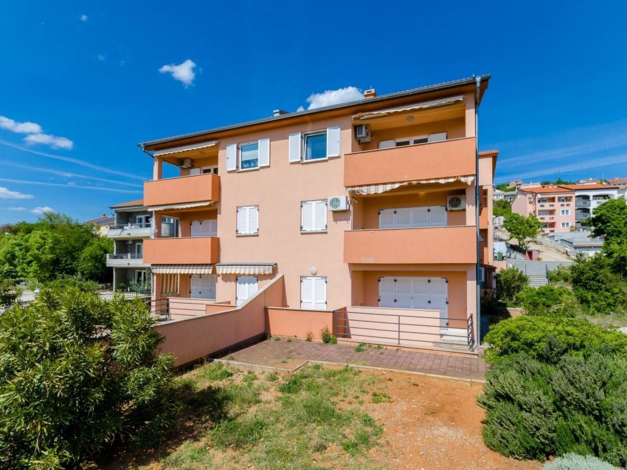 Un immeuble résidentiel à plusieurs étages, doté de balcons, d’une cour pavée et entouré de verdure.