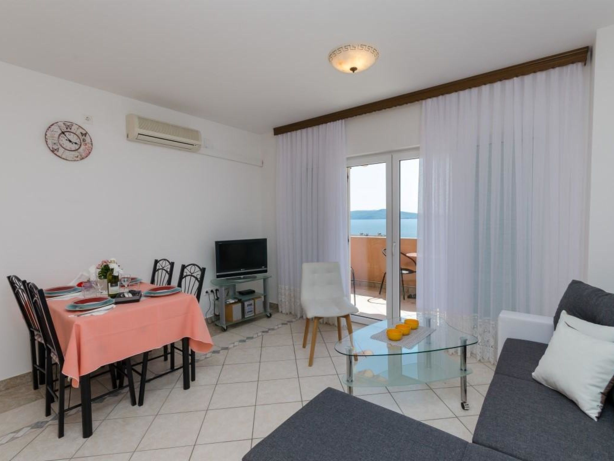 A living room with a dining area, a sofa, a glass coffee table, a TV, and a view of the sea through a sliding glass door.