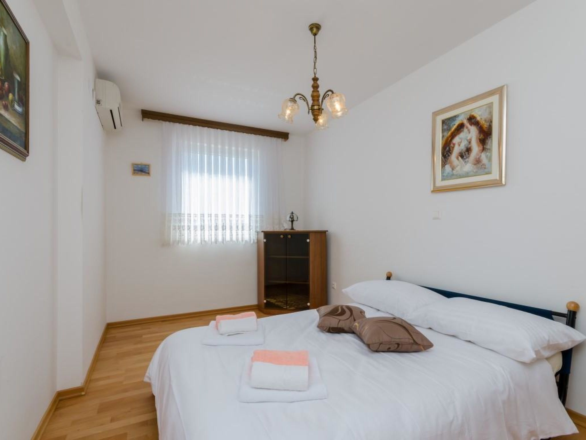 A bedroom with a bed, a dresser, a window with curtains, a chandelier, and a painting on the wall.
