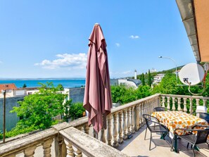 Ein Balkon mit Tisch und Stühlen, einem pinken Sonnenschirm und Blick auf das Meer und Gebäude.