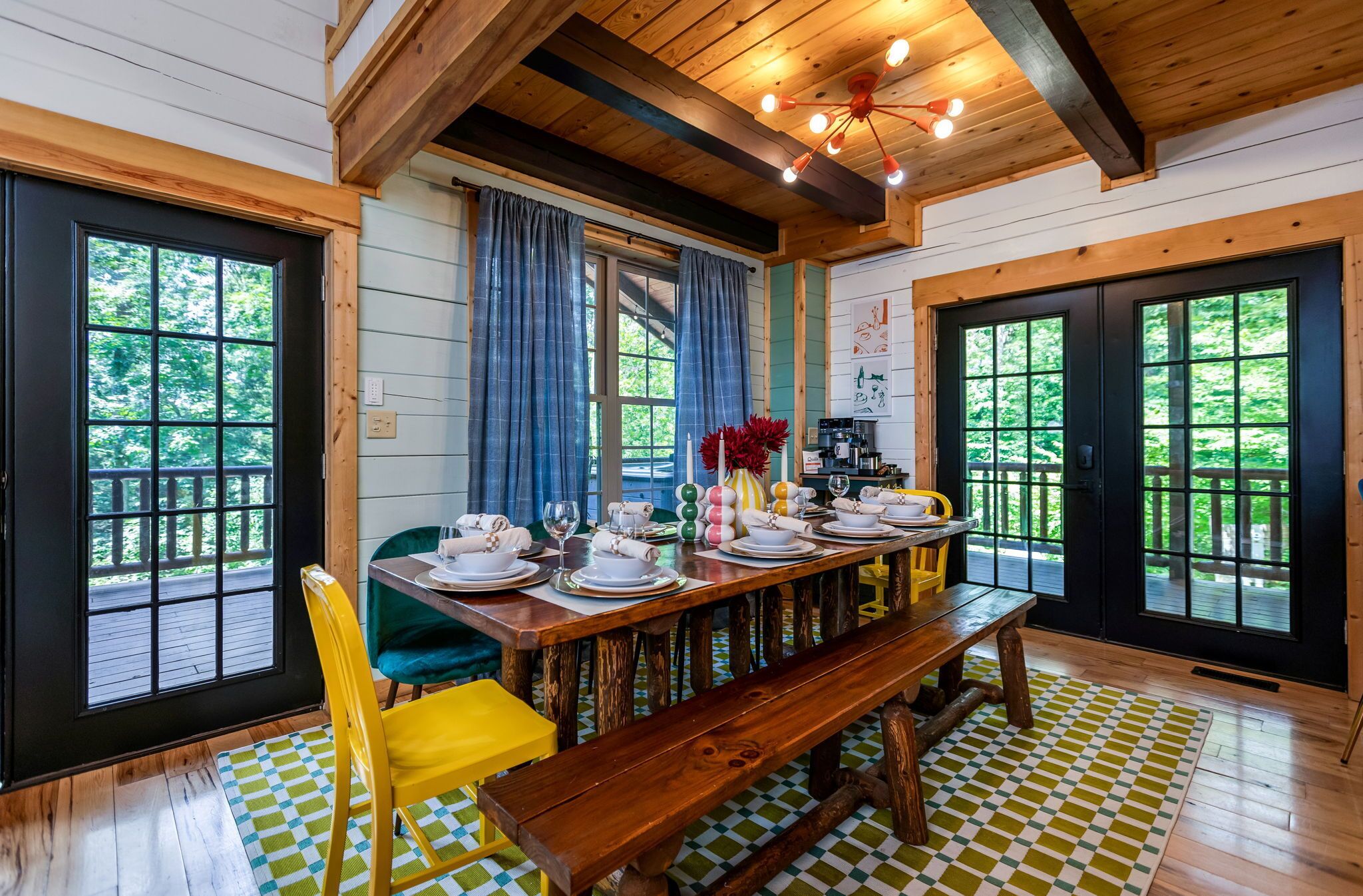 A dining area with a wooden table set for four, a bench, and a view of the outdoors through large windows.