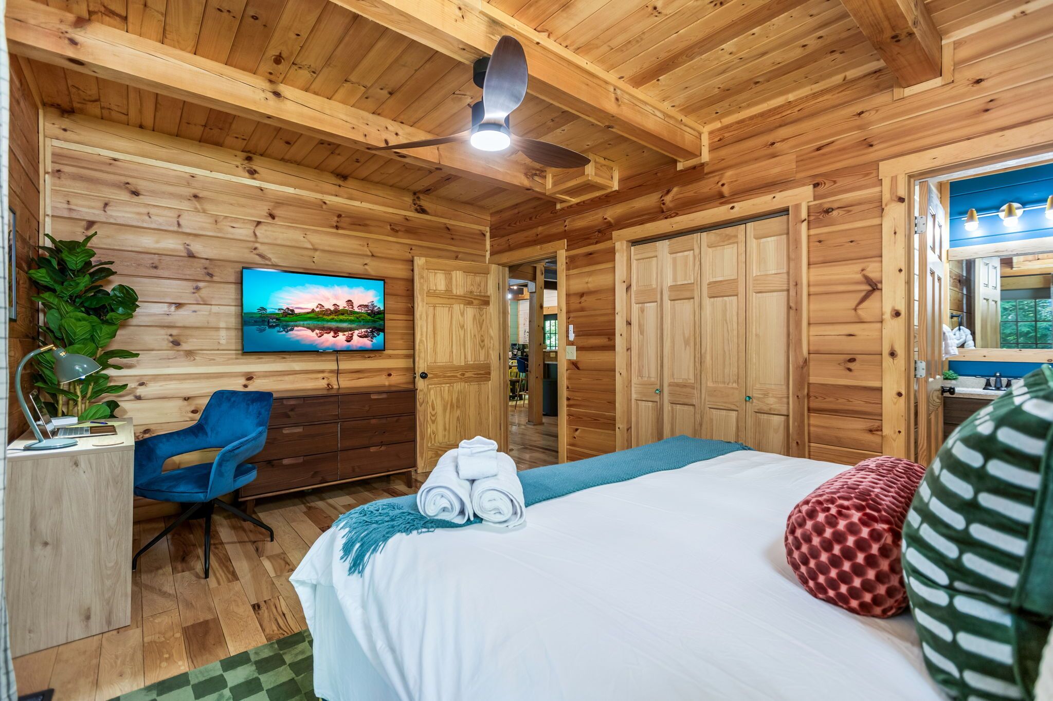 A wooden cabin-style room with a bed, a desk, a chair, a TV, and a ceiling fan.