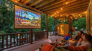Eine Holzterrasse mit einer großen Leinwand, Sitzgelegenheiten im Freien und einem rustikalen Gebäude im Hintergrund.