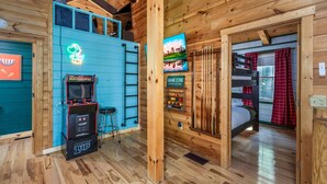 Interior de una cabaña de madera con un letrero de neón, máquinas de arcade, una cama litera y un televisor.