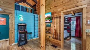 A wooden cabin interior with a neon sign, arcade machines, a bunk bed, and a TV.