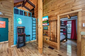 Interior de uma cabana de madeira com um letreiro de neon, máquinas de jogos eletrônicos, uma cama beliche e uma TV.