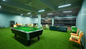 A pool table with a green felt surface, surrounded by chairs and tables in a room with green carpet and light blue walls. - Ikshana Resort and Spa (Mawal)