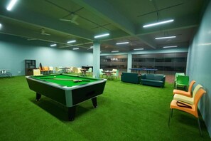 A pool table with a green felt surface, surrounded by chairs and tables in a room with green carpet and light blue walls. - Ikshana Resort and Spa (Mawal)