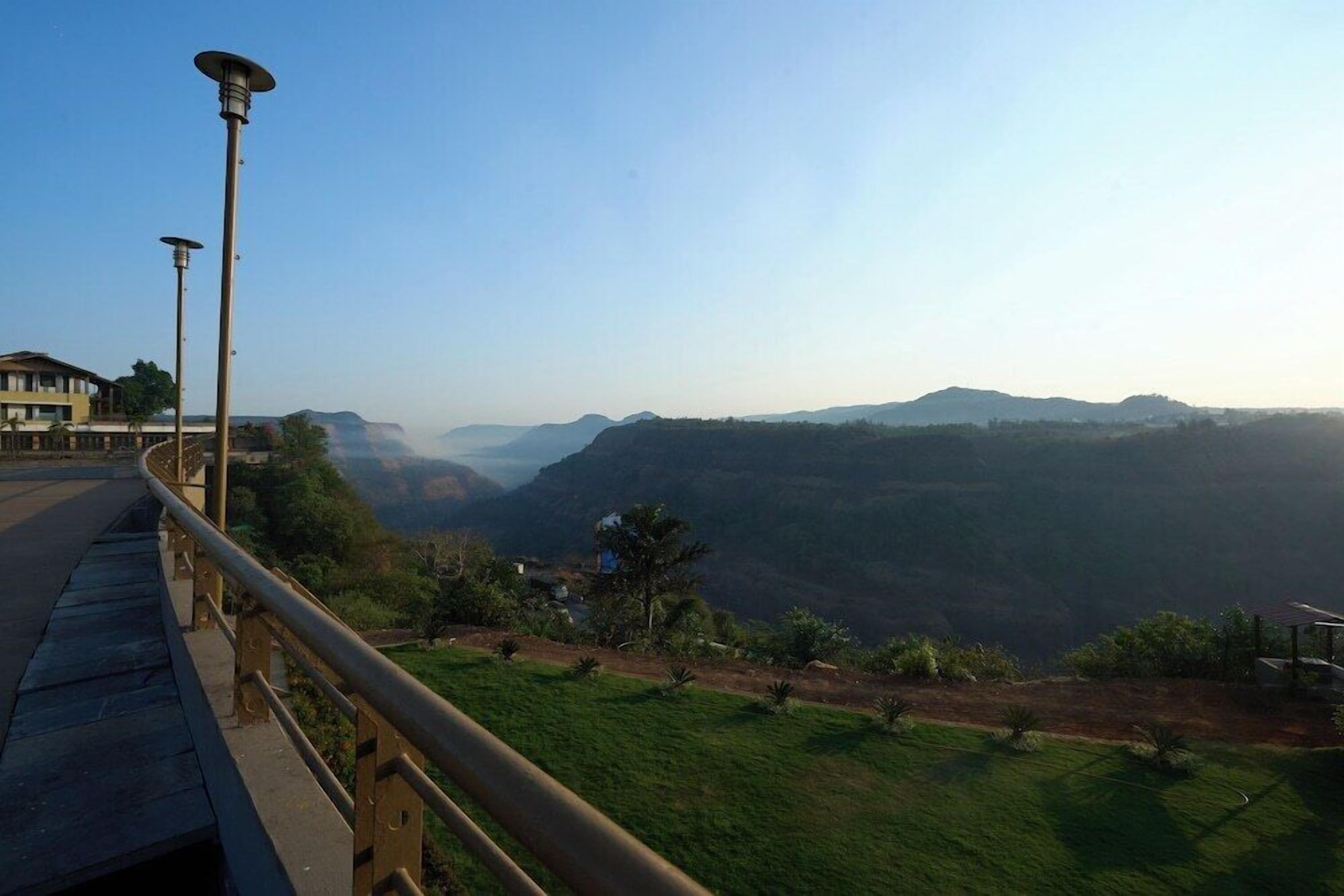 A road with a guardrail, a lamp post, and a view of a valley with distant mountains.