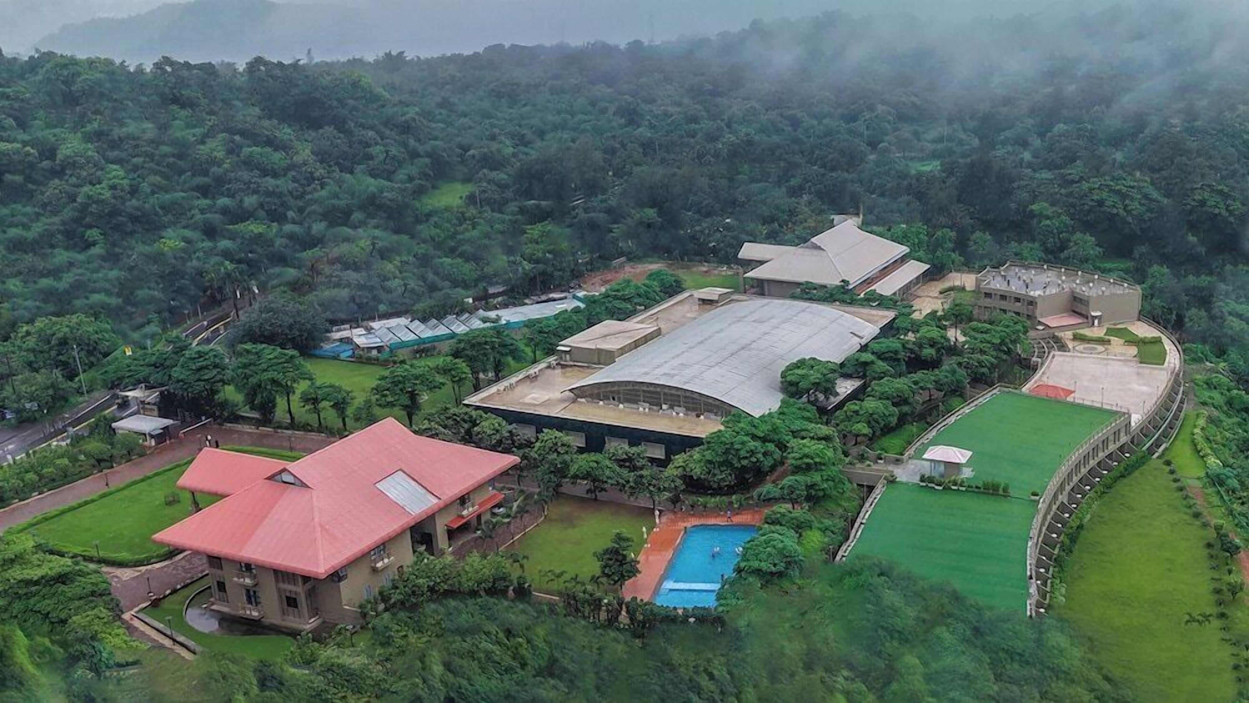 Vista aérea de um grande complexo de edifícios modernos, rodeado por vegetação exuberante, com piscina e quadra de tênis visíveis.