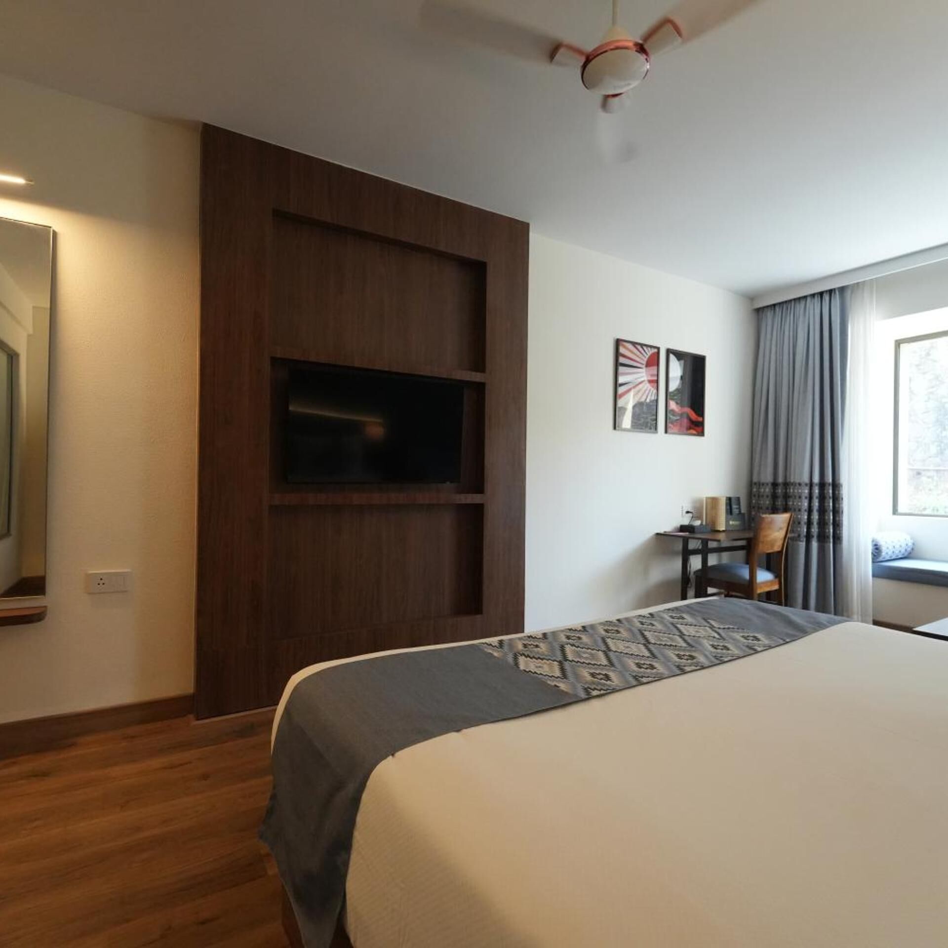 Habitación de hotel con cama, televisor montado en un panel de madera, escritorio con silla y ventana con cortinas.