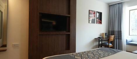 Habitación de hotel con cama, televisor montado en un panel de madera, escritorio con silla y ventana con cortinas.