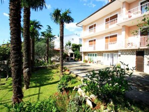 Un edificio a due piani con balconi, facciata in pietra e un giardino curato con palme e arbusti.
