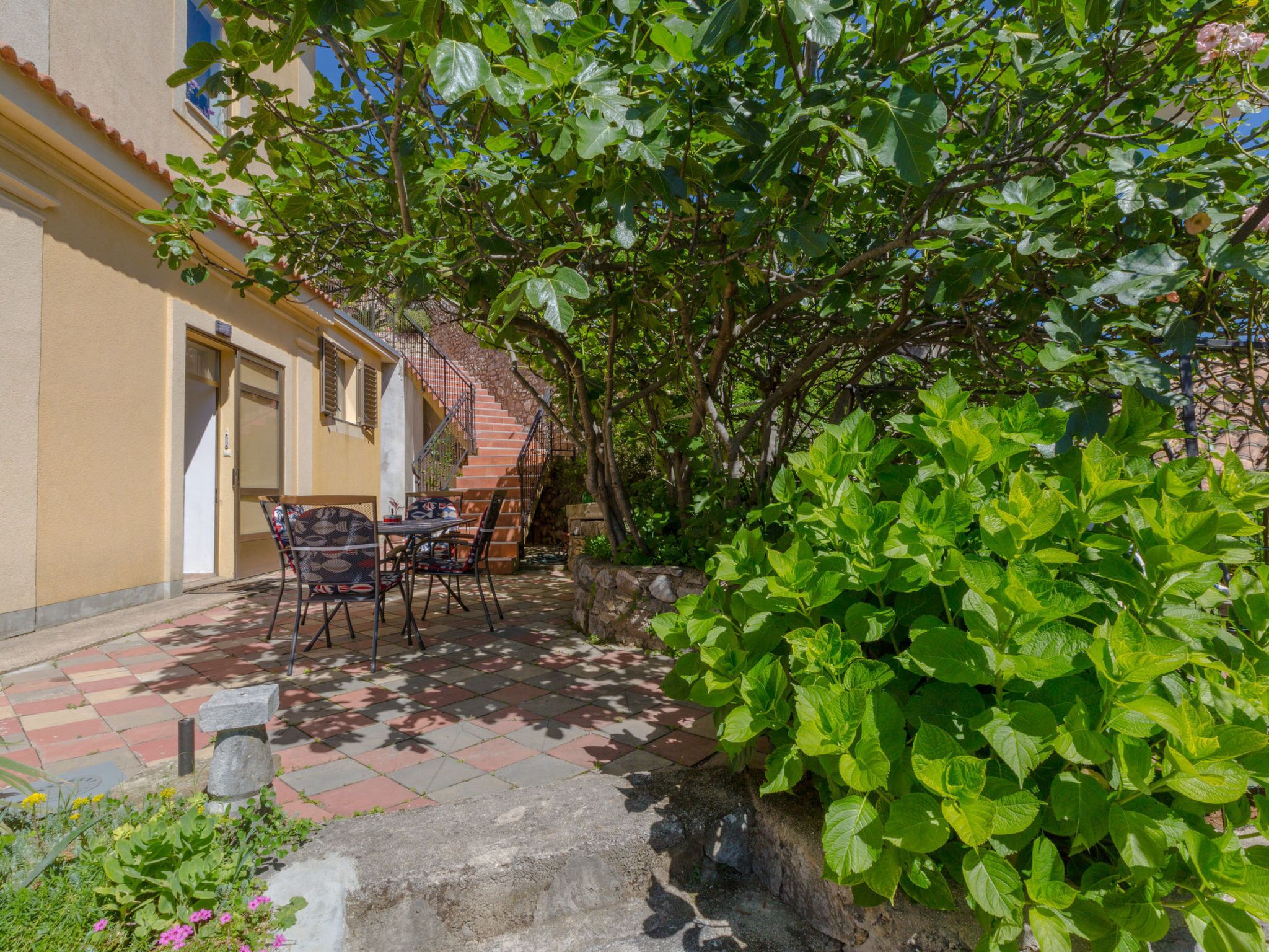A courtyard with a table and chairs, a staircase, and a garden with green foliage.