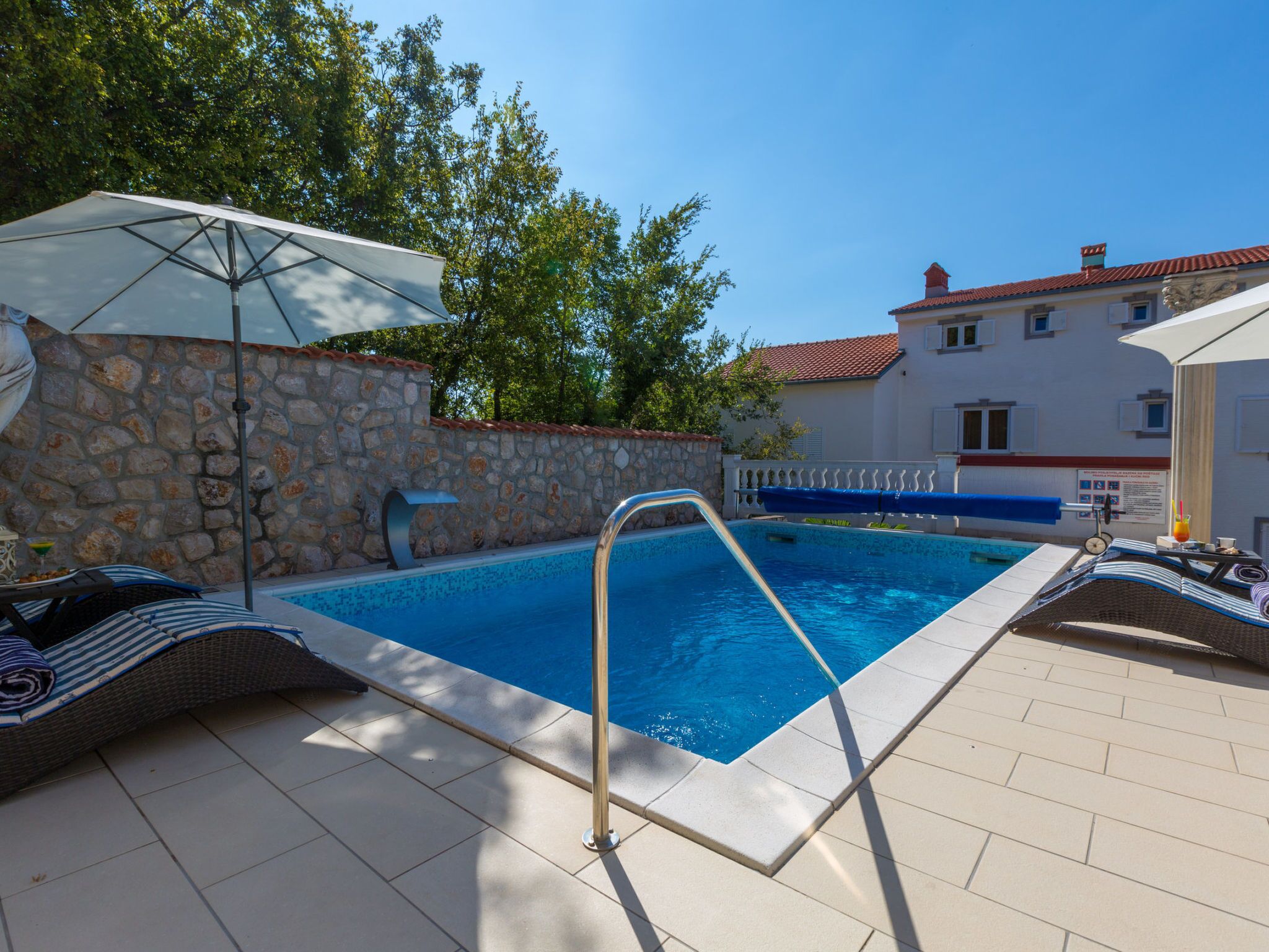 A swimming pool with a stone wall, lounge chairs, and an umbrella.