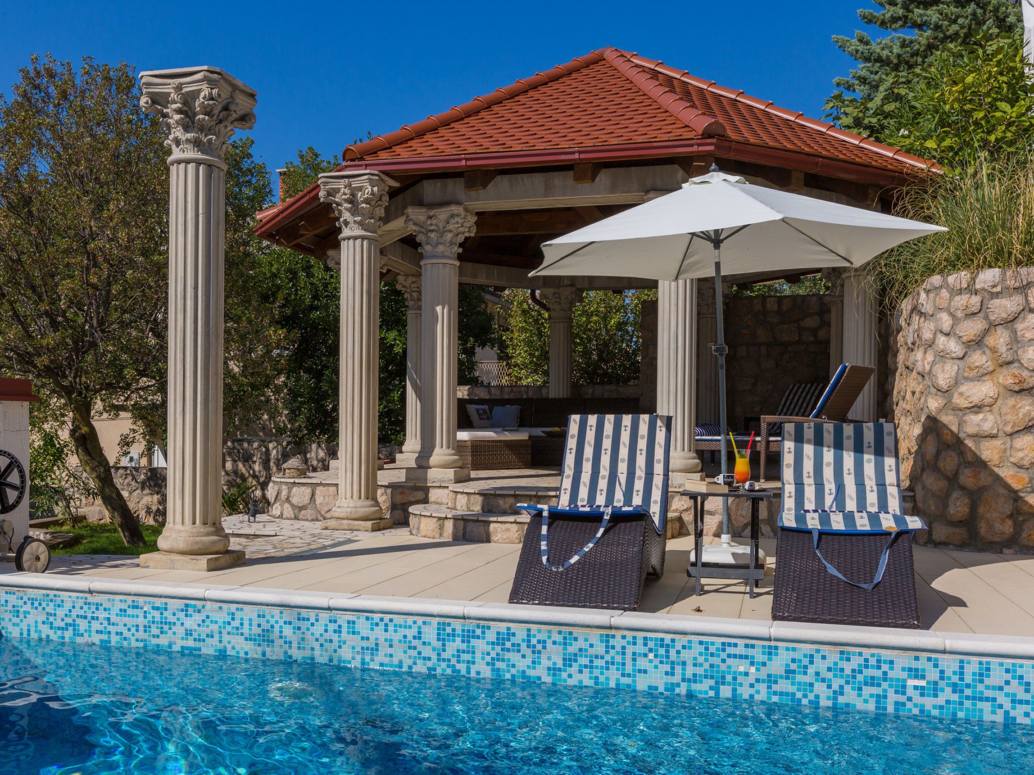 A pool area with a gazebo, lounge chairs, and a stone wall.