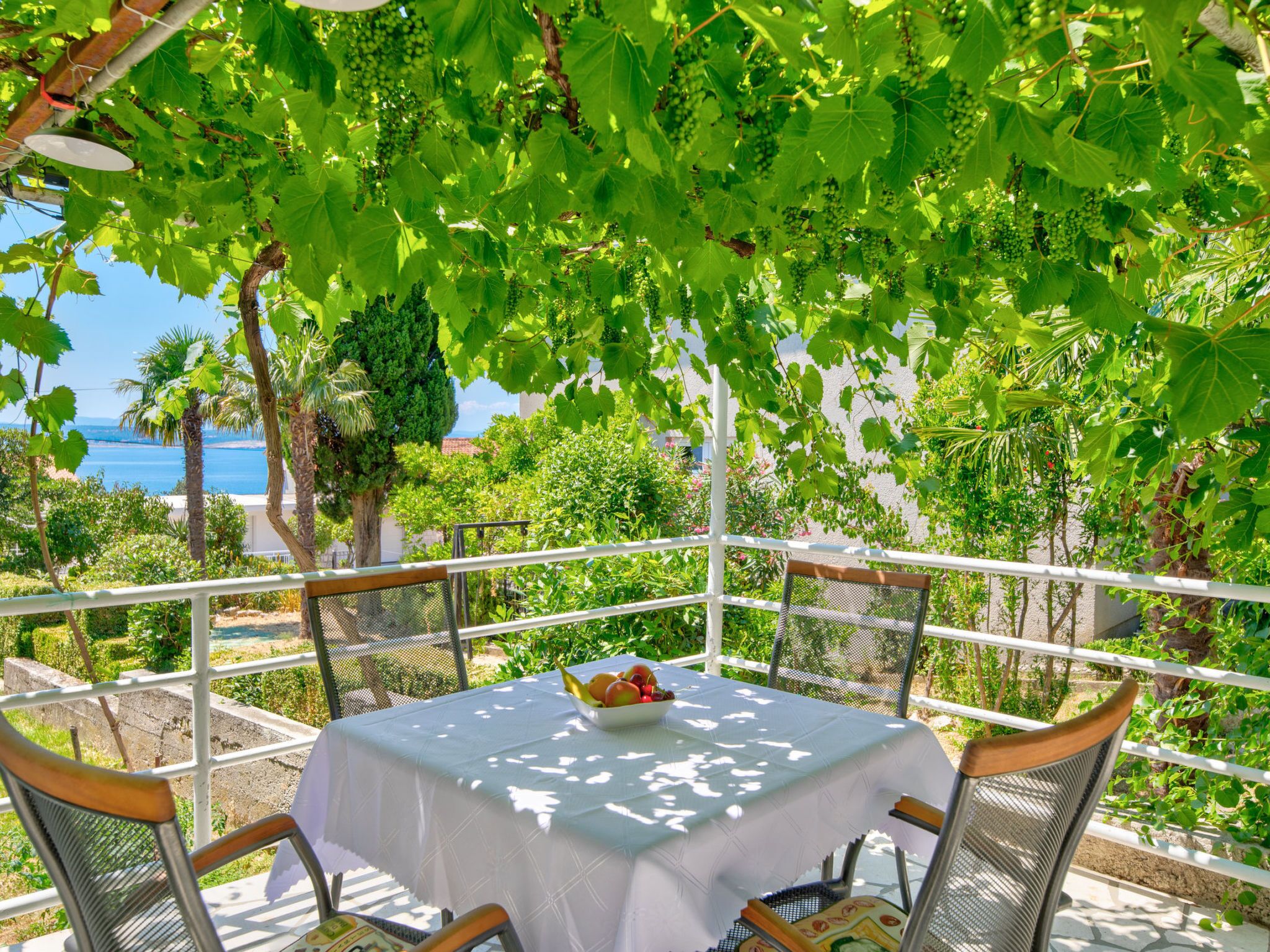A table with a white tablecloth, surrounded by metal chairs, under a pergola with green foliage.
