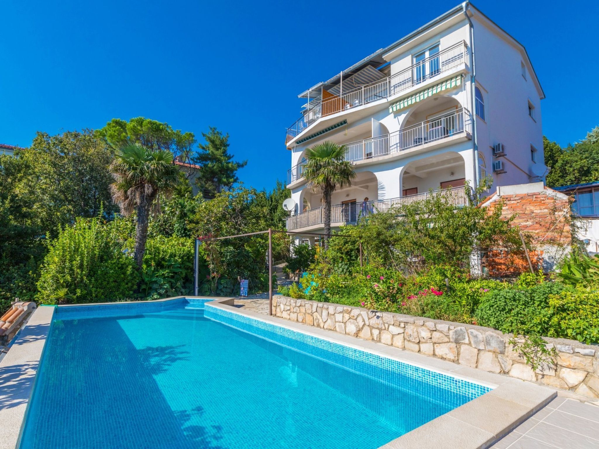 A two-story building with a swimming pool, surrounded by greenery and a clear blue sky.