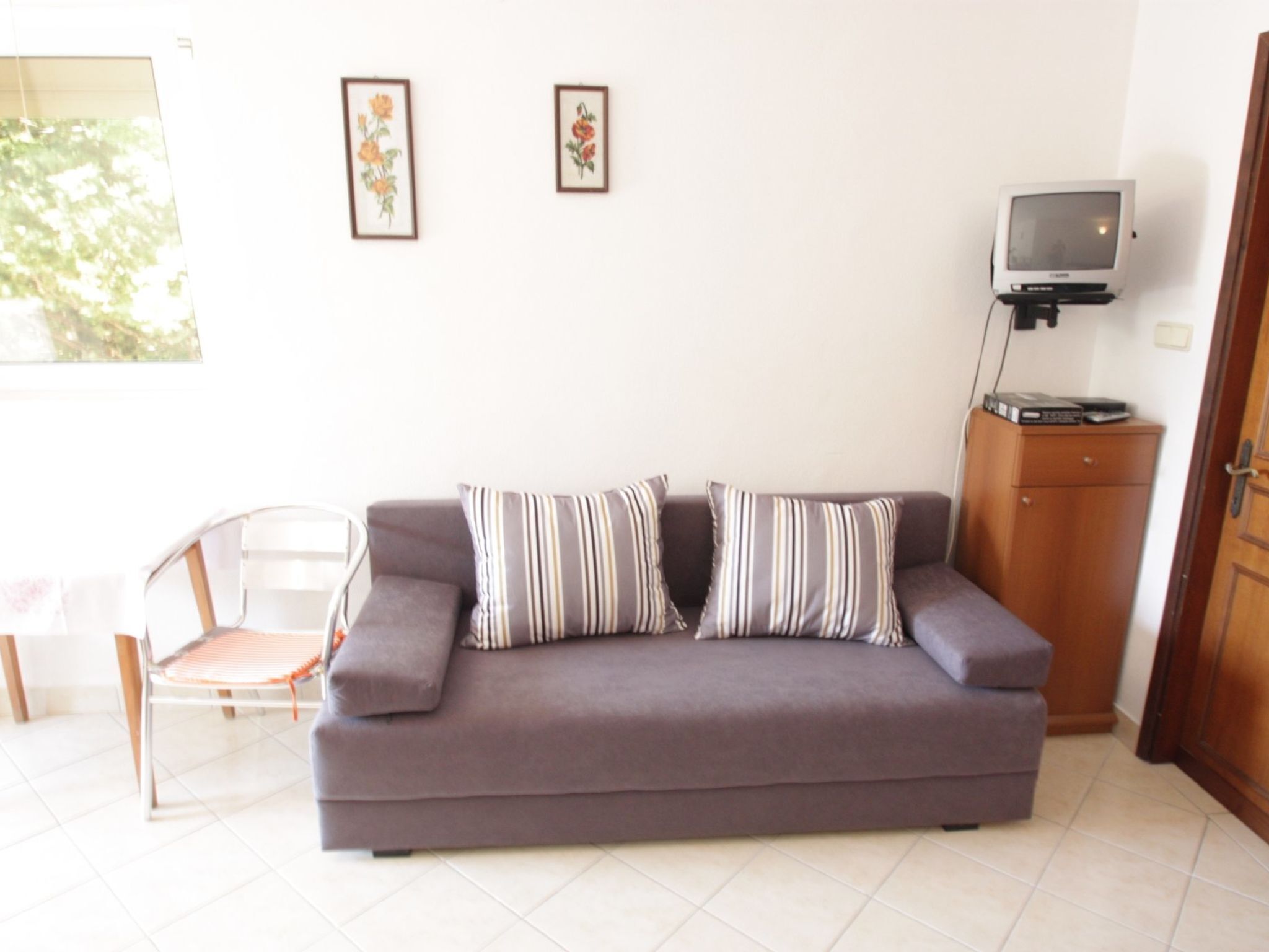 A living room with a sofa, two framed floral pictures on the wall, a television on a wooden cabinet, and a chair with a striped cushion.