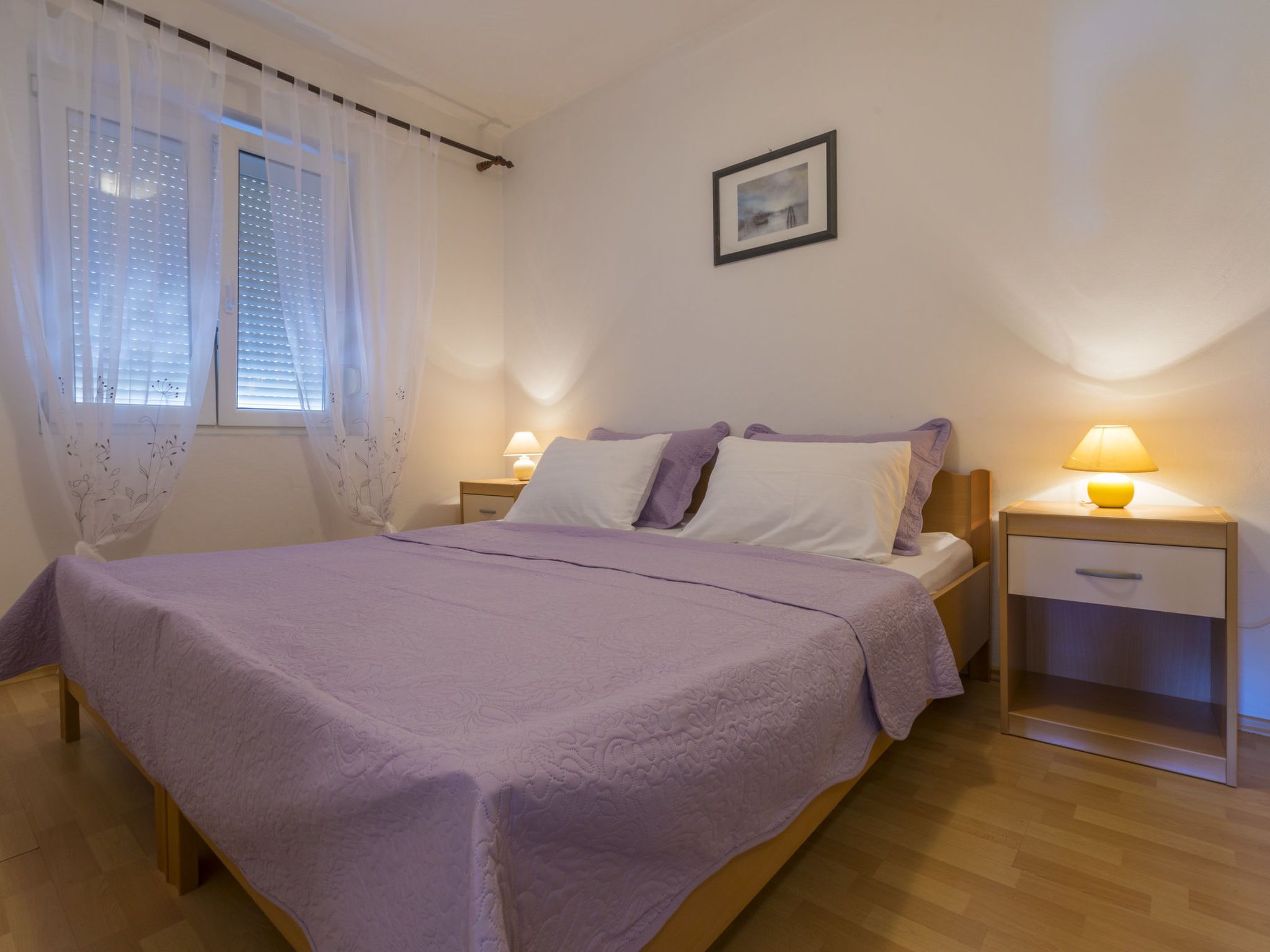 A bedroom with a bed, two bedside tables, a nightstand, a window with blinds, and a framed picture on the wall.