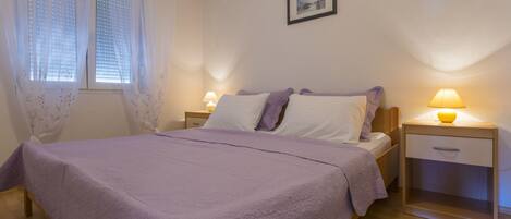 A bedroom with a bed, two bedside tables, a nightstand, a window with blinds, and a framed picture on the wall.