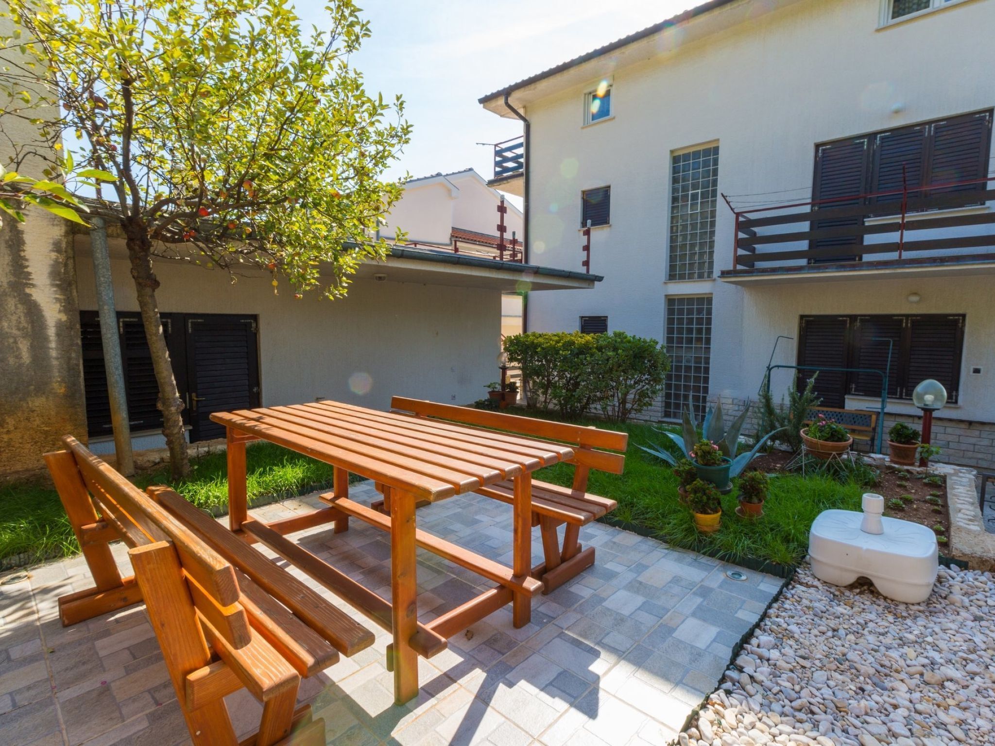 A courtyard with a wooden table and benches, surrounded by greenery and a building with balconies.