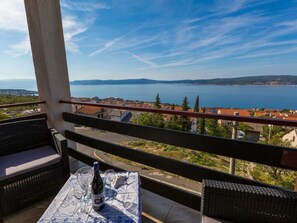 En balkon med fletmøbler og et bord dækket til to, med udsigt over en kystby og et stort vandområde.