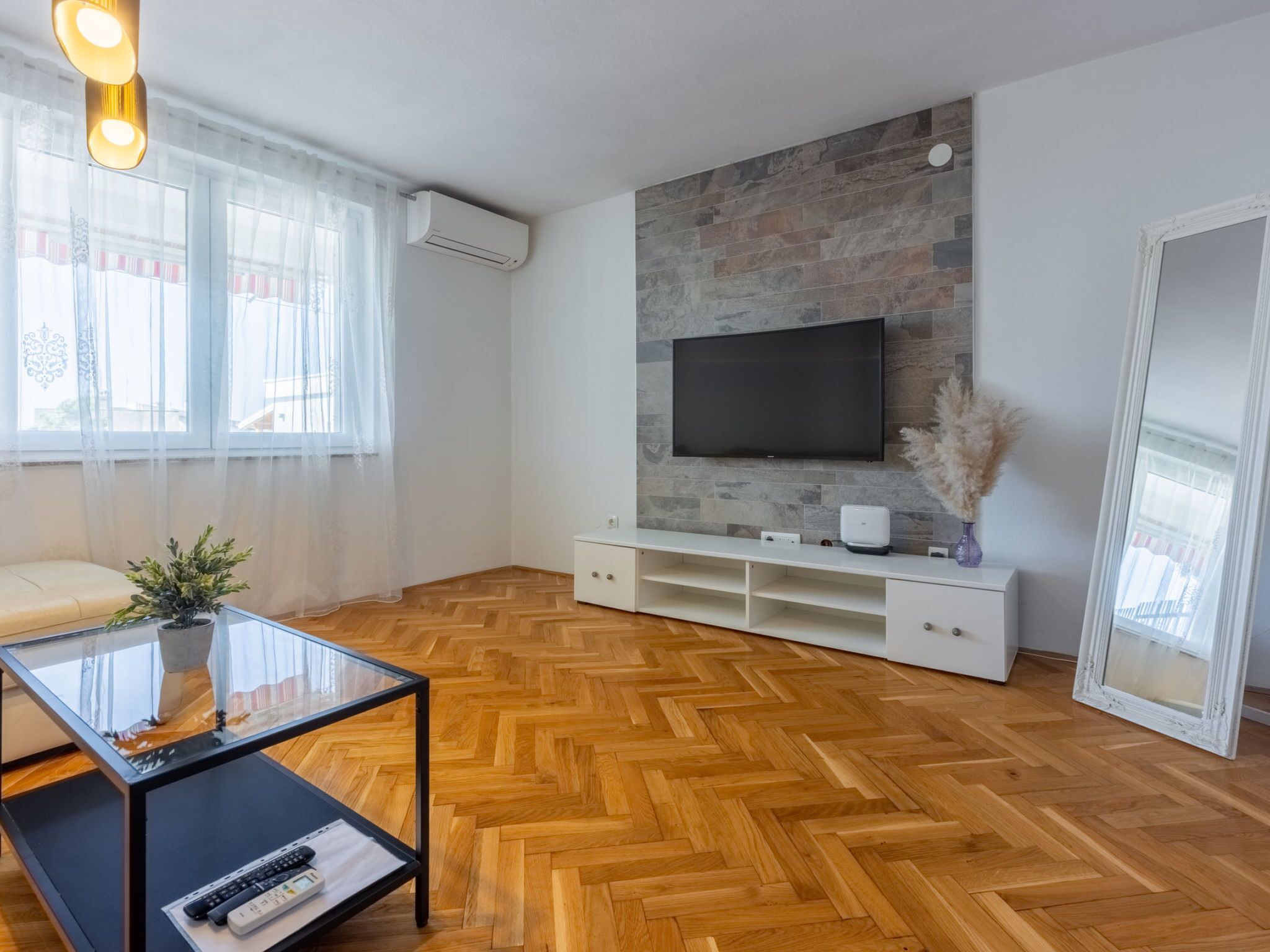 A modern living room with a herringbone wooden floor, a flat-screen TV mounted on a stone wall, a glass coffee table, and a white entertainment unit.