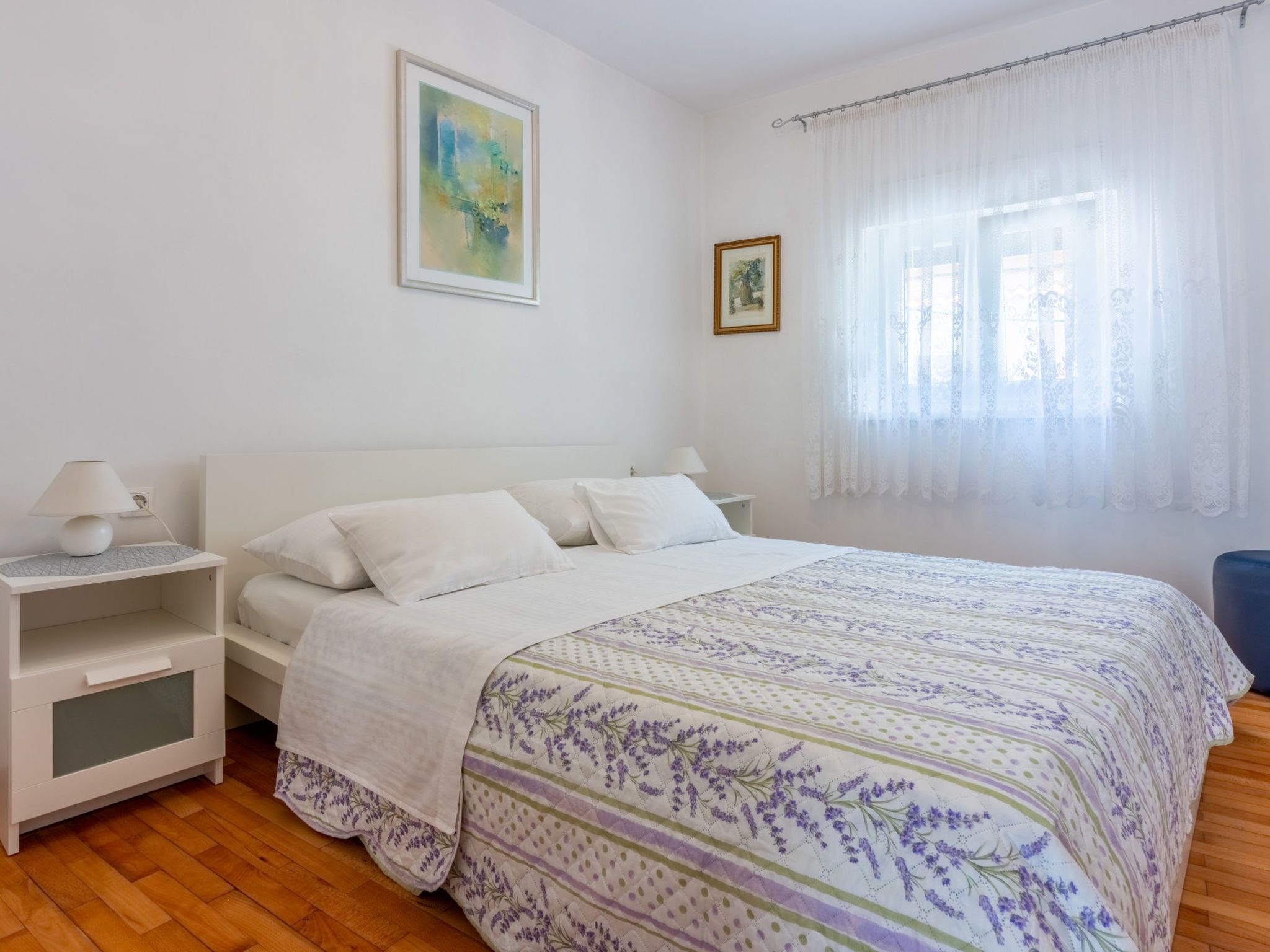 A bedroom with a bed, nightstands, a window with sheer curtains, and a painting on the wall.