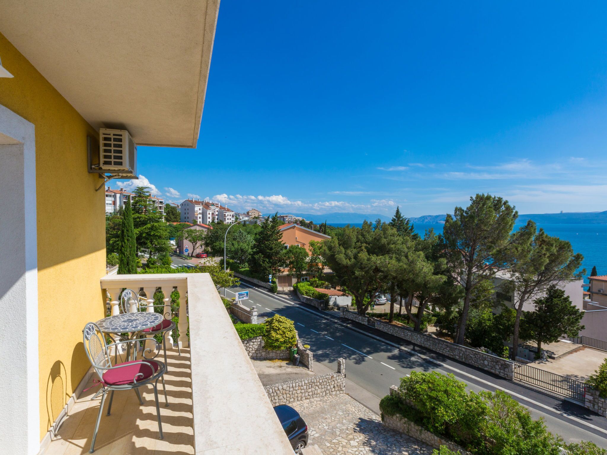 A balcony with a table and chairs, overlooking a road and buildings, with a clear view of the sea and mountains in the distance.