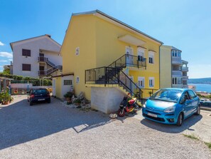 Ein gelbes Gebäude mit Treppe, ein parkendes blaues Auto davor und im Hintergrund der Blick aufs Meer.