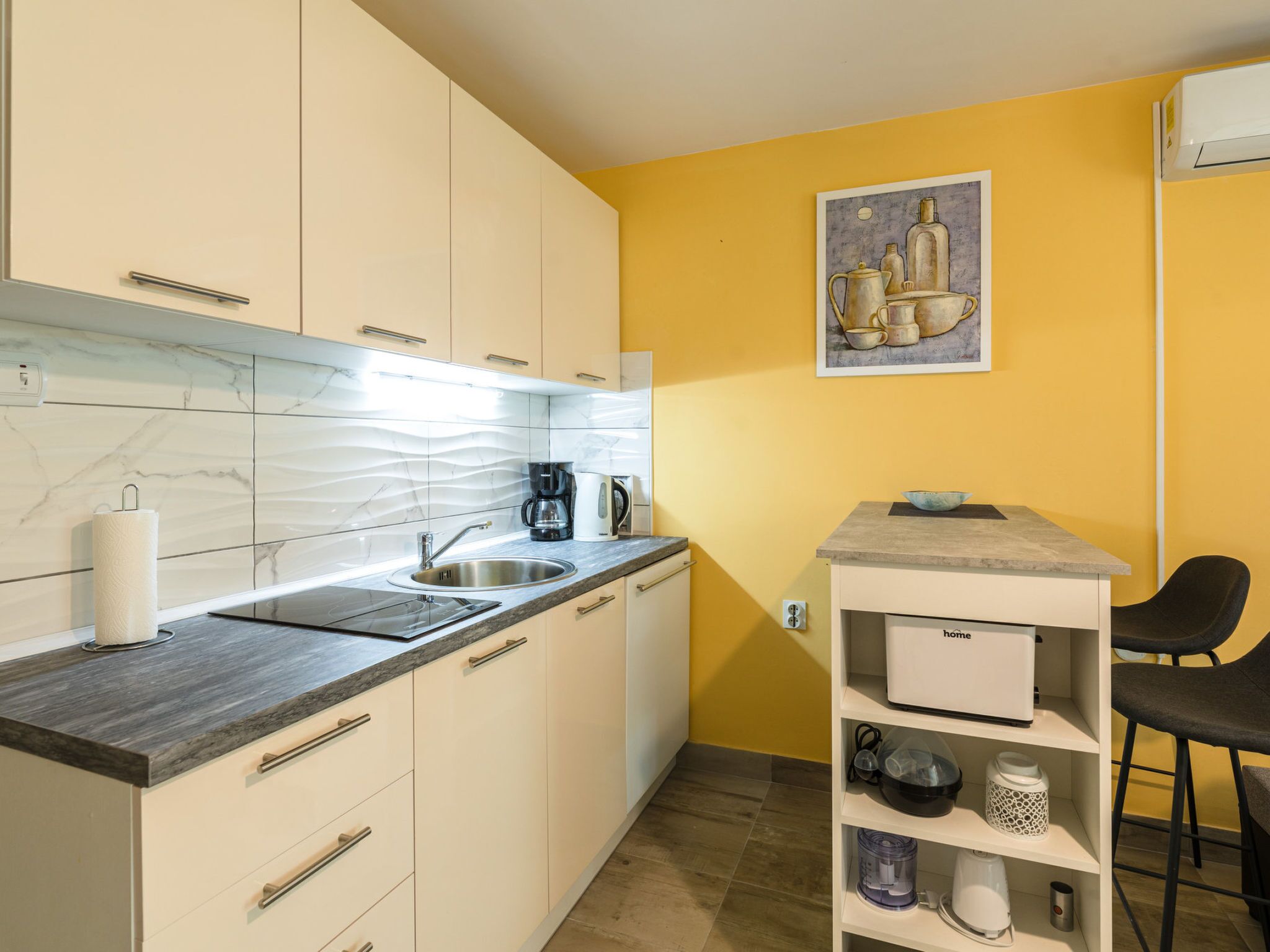 Cocina compacta con armarios blancos, salpicadero de mármol, cafetera y un pequeño comedor con una silla.