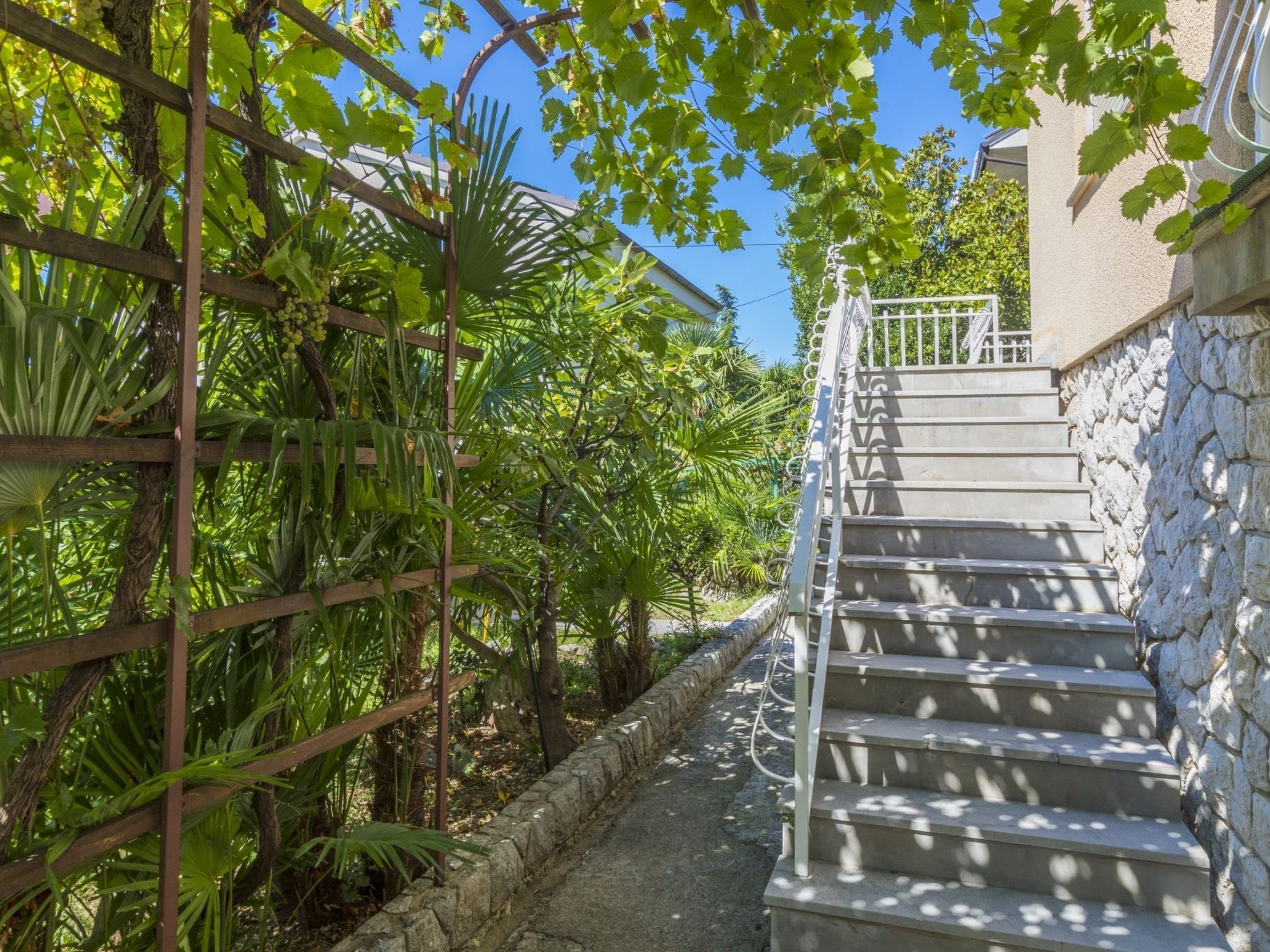 A staircase with stone walls and a metal railing, surrounded by lush greenery and a clear blue sky.