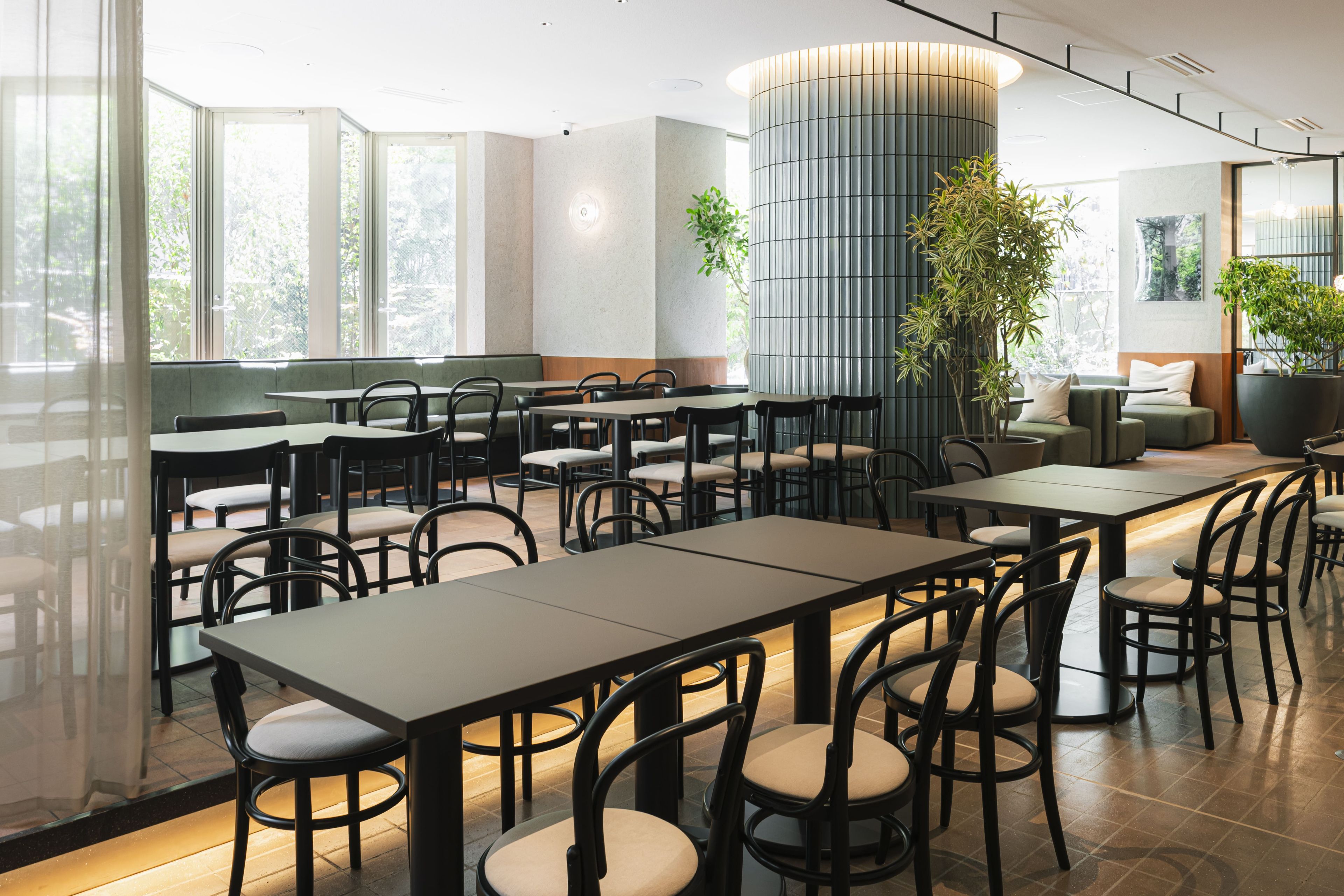 A modern restaurant with multiple tables and chairs, large windows, and indoor plants.