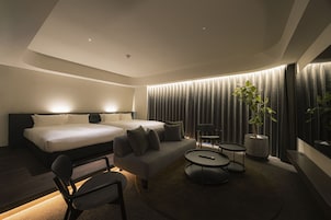 A modern hotel room with a large bed, a sofa, two small tables, a chair, and a potted plant.