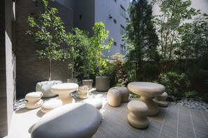 Área externa com mesas de pedra e vasos de plantas, cercada por edifícios e árvores.