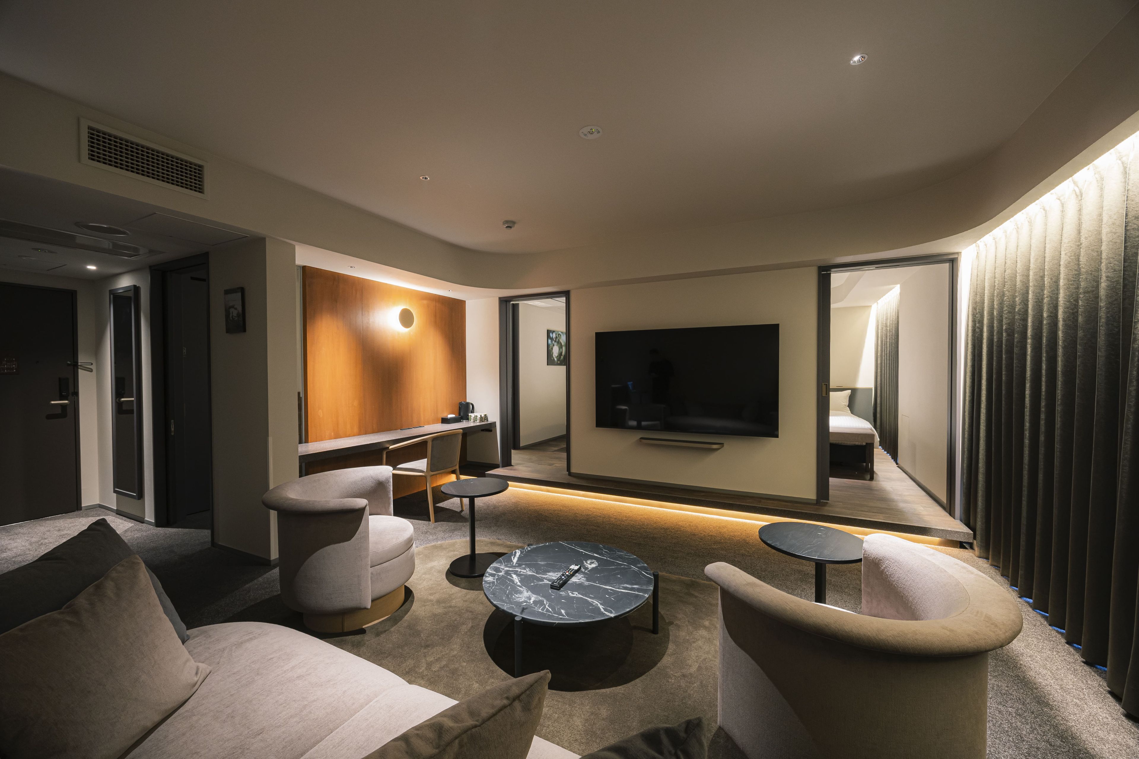 A modern hotel room with a sofa, armchairs, a coffee table, a TV, and a large mirror.