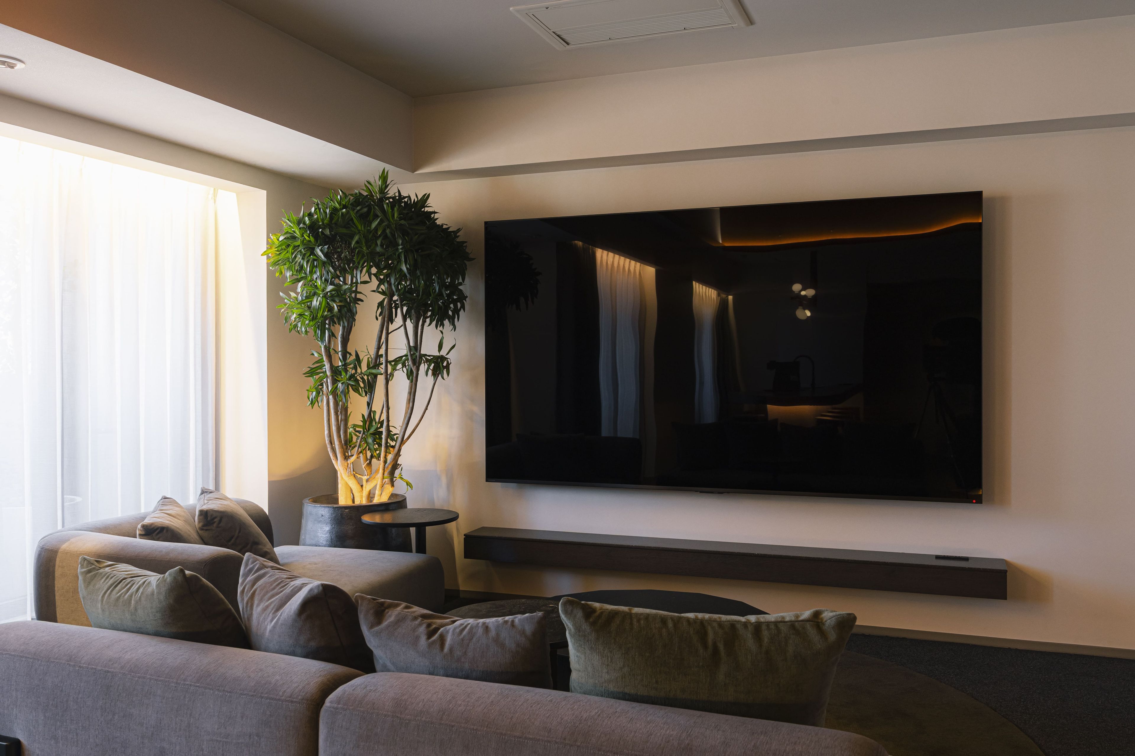 Una sala de estar moderna con un televisor de pantalla plana grande montado en la pared, un sofá gris con varios cojines y una planta en maceta.