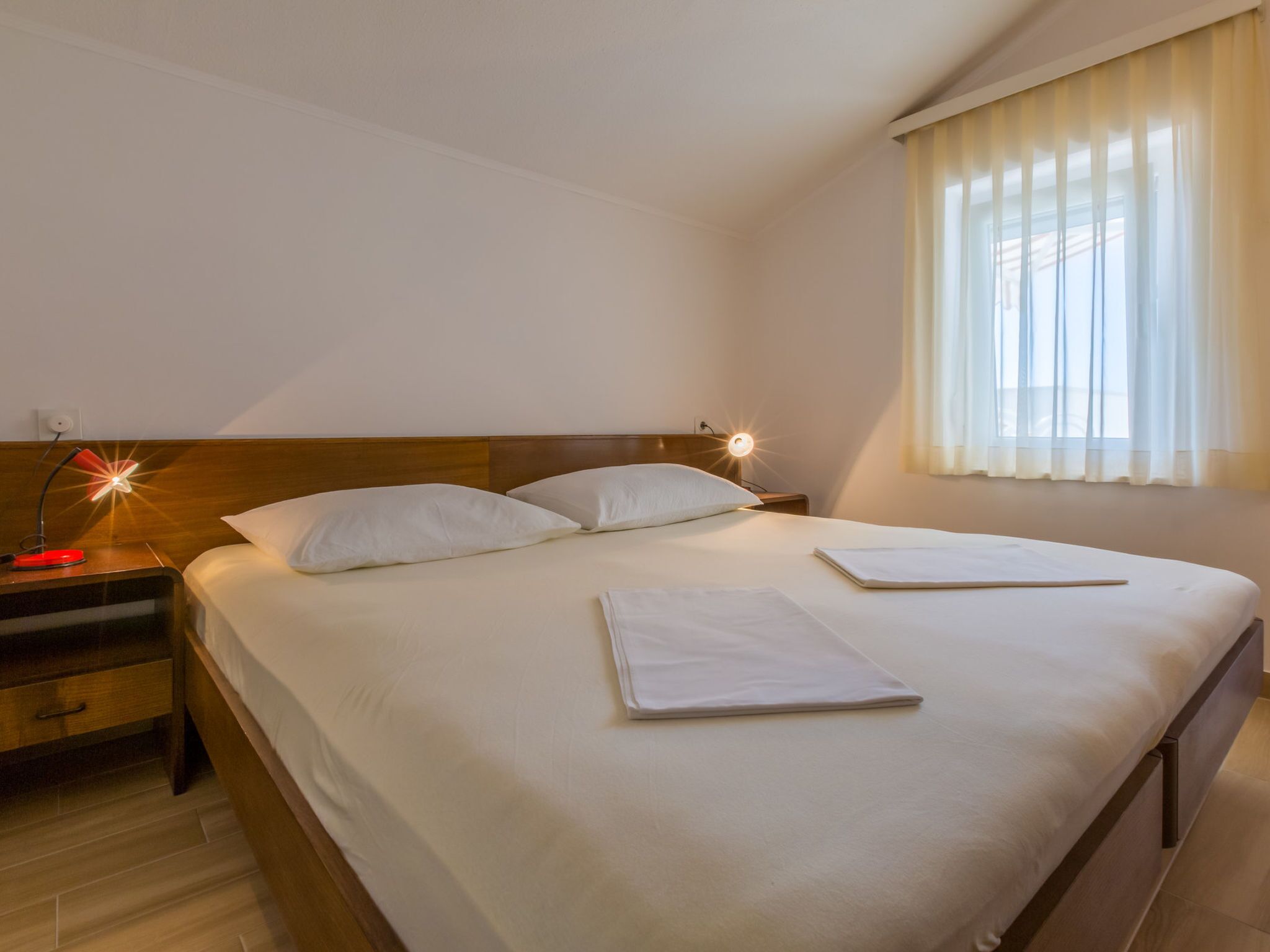 A neatly made bed with white linens, a bedside lamp, and a window with sheer curtains.